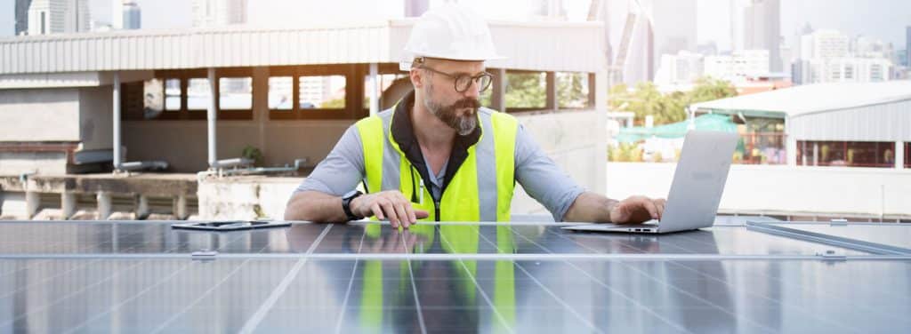 Servicetechniker mit Helm und Warnweste prüft eine Photovoltaikanlage auf einem Dach mithilfe eines Laptops.