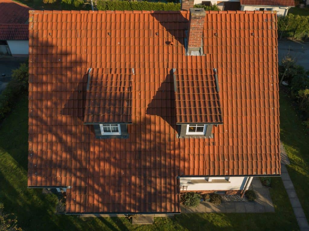 Senkrechte Luftaufnahme eines Ziegeldachs mit zwei Gauben und Schornstein, auf dem der Schatten eines Baums liegt.